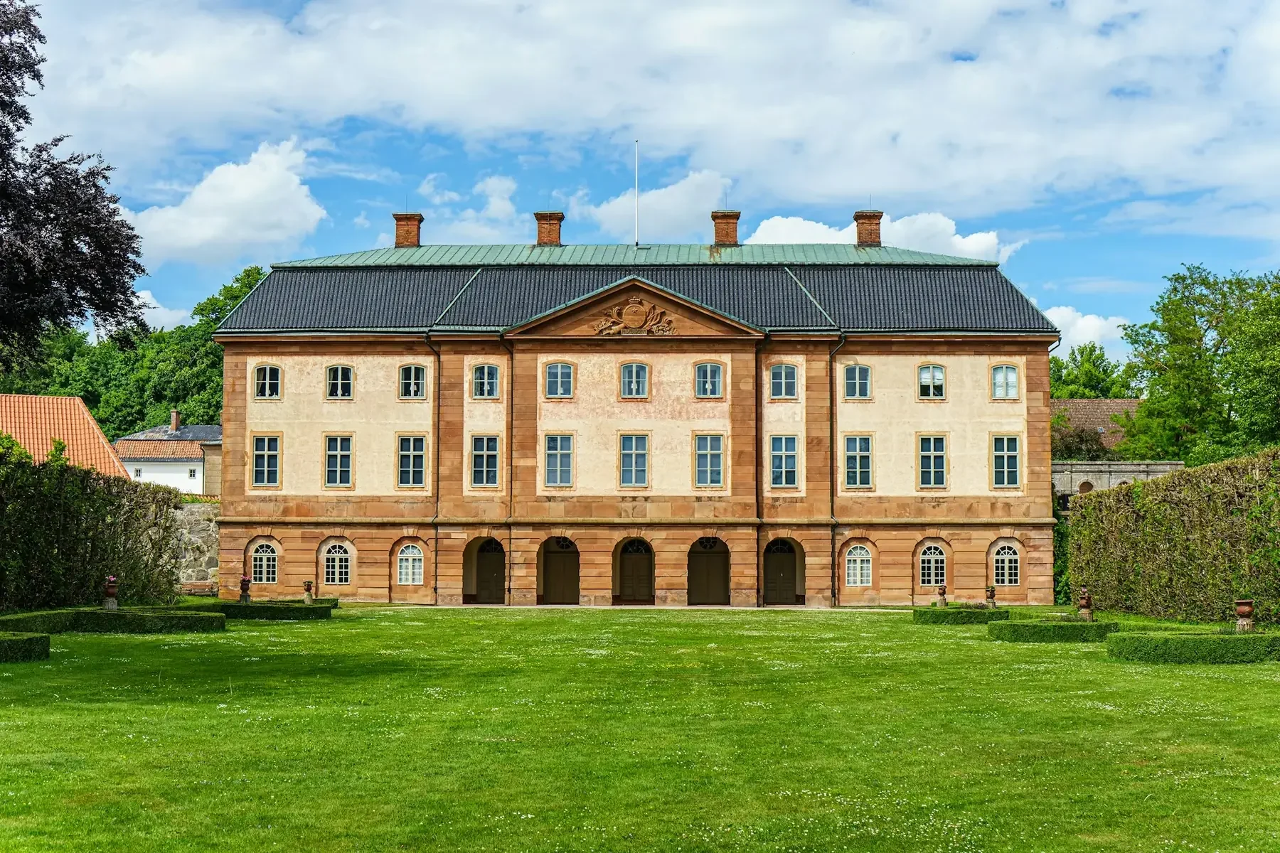 Ståtlig herrgård i ljus puts och tegel framför en stor gräsmatta en sommardag.