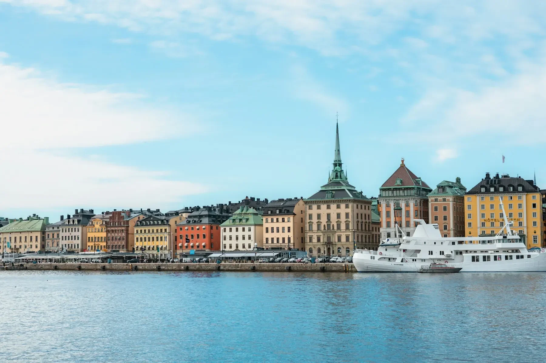 Färgglada hus längs vattnet i Gamla stan i Stockholm, med ett vitt passagerarfartyg vid kajen.