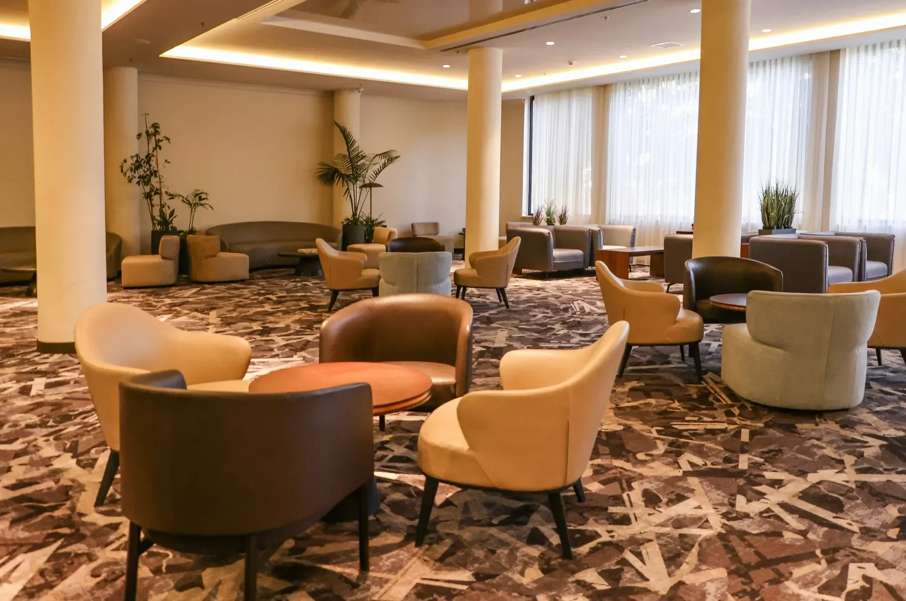 Rymlig hotellounge med fåtöljer, små bord, pelare och stora fönster med gardiner.