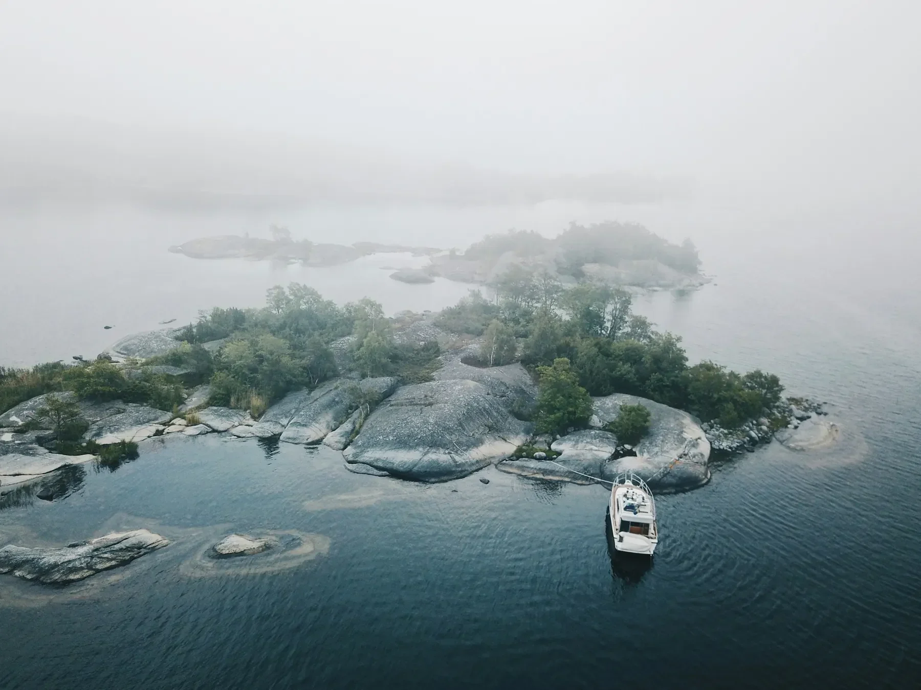 Flygbild över en skärgårdsö med släta hällar och tallar i morgondis, med en vit motorbåt bredvid.