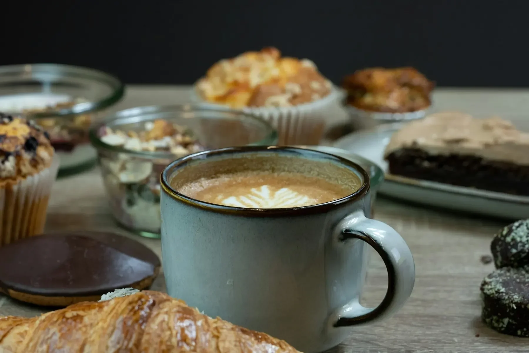 Kaffe med mjölkskum i en keramikkopp omgiven av kakor, bullar och en croissant på ett bord.